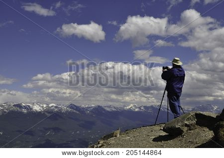 Man with his camera on a tripod at the top of a ridge photographing the mountain range across from him.