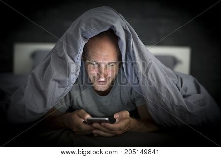 A young cell phone addict man awake late at night in bed using smartphone
