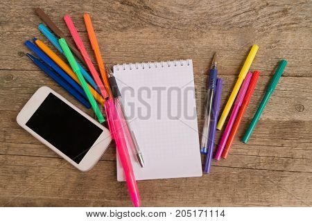 Open white notepad with colorful felt-tip pens and ball pens with smartphone on the wooden table. Space for text