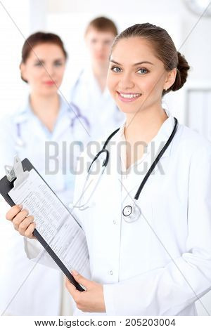 Happy female doctor keeping medical clipboard while medical staff are at the background. Successful team at health care and medicine concept
