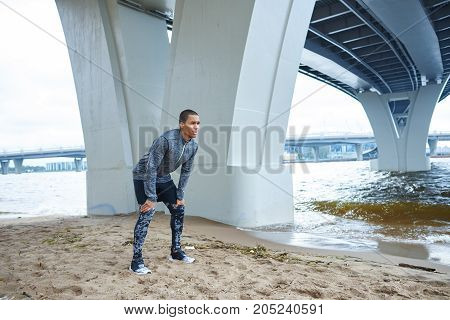 Sports fitness people and healthy lifestyle. Muscular athletic dark-skinned male runner in sportswear catching breath after cardio workout standing on sand with hands on knees and leaning forward