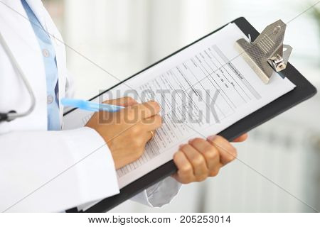 Close-up of a female doctor filling  up medical form at clipboard while standing straight in hospital.