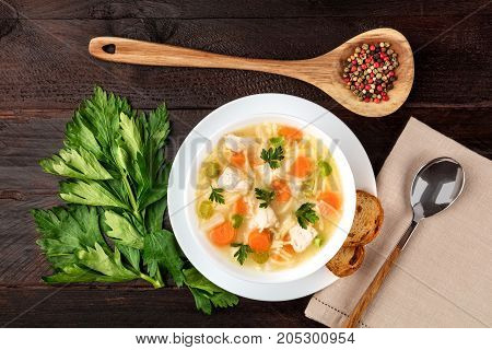 An overhead photo of a plate of chicken and noodles soup, shot from above on a dark rustic texture with a spoon, a wooden ladle with peppercorns, slices of bread, a celery branch, and a place for text