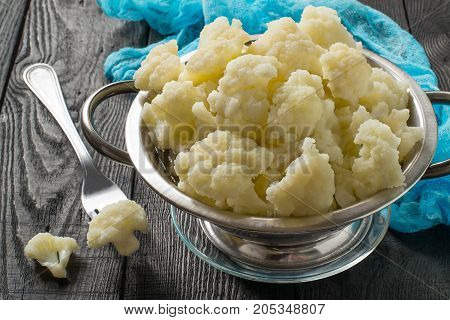 Blanched cauliflower in colander on gray wooden background. White cauliflower inflorescences in metal colander blue gauze napkin on table. Preparing for diet dinner