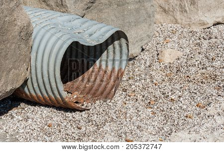 Old rusted and dented corrugated metal pipe emerging from between rocks onto shell-covered beach.