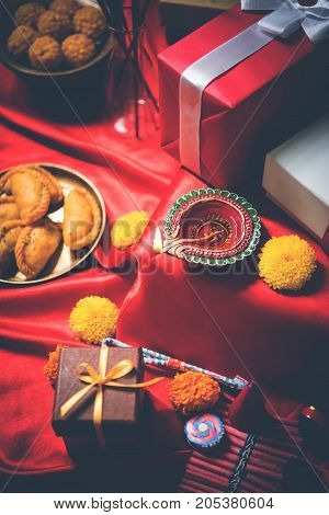 stock photo showing Diwali still life with Diya or oil Lamp, gift boxes, sweets and fire crackers with flowers arranged over red satin cloth with bokeh