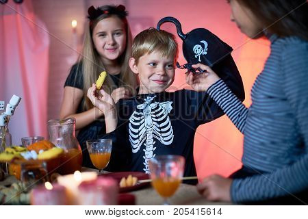 Girl showing toy scorpion to smiling cute boy in skeleton costume with hook instead of hand eating cookie at Halloween party, cheerful pretty girl behind him looking at camera