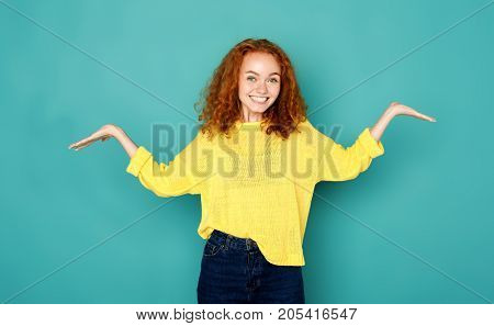 Young smiling puzzled woman with arms out. Asking what is problem, who cares, so what, I don't know on blue background.