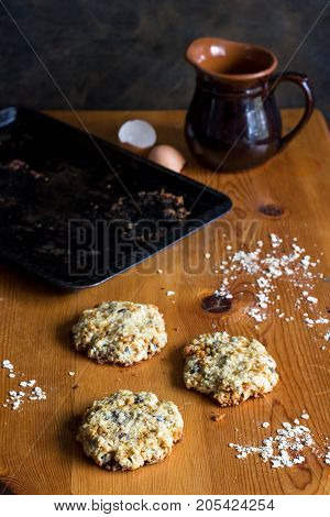 Home baked oats and date cookies on rustic wooden table with egg shell jug and baking tin in background. Vertical photo with copy space