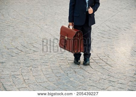 Close up shot of future businessman holding leather briefcase while staying on pavage street outdoor in city. Braun leather bag with golden belt on gray stone background. Copy space available.