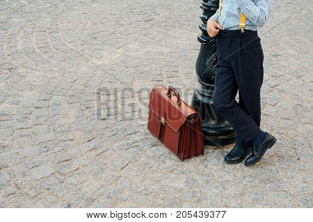 Close up shot, future businessman leather briefcase while staying with crossed legs on pavage street outdoor in city. Braun leather bag with golden belt on gray stone background. Copy space available.