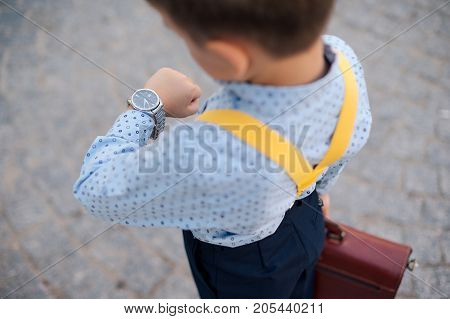 Concept future businessman. Boy portrait in dark blue pants, baby blue shirt and yellow suspenders, standing on pedestrian street with braun briefcase looking to his watch, urban background.