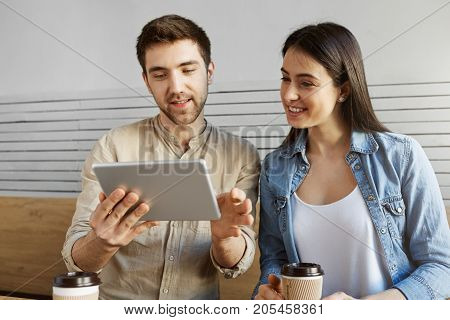 Pair of young enterpreners sitting in cafe working on new startup project. Guy showing website statistics on digital tablet