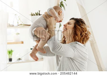 Happy smiling mother playing with newborn child in comfy light bedroom in front of window. Moments of motherhood happiness with kids. Family concept