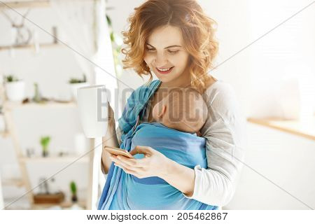 Cheerful smiling mother drinks cocoa and texting her beloved husband while small son dozing in baby slim. Atmosphere of love and protection