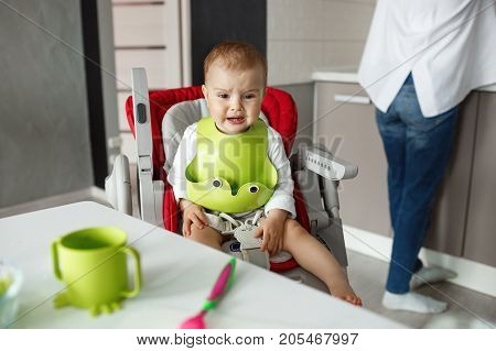 Portrait of little scared baby boy sitting in baby chair in kitchen, crying and screaming while mother cooking him food