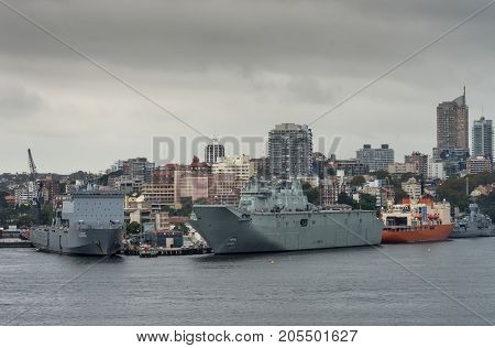 Sydney Australia - March 21 2017: Several Navy vessels docked along Garden Island quays under rainy stormy cloudscape and city skyline. People and activity on deck.