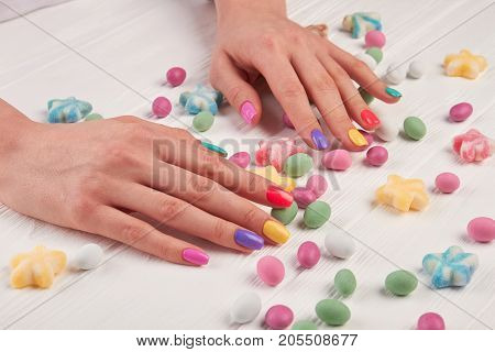 Girls manicured hands and sweets. Girls hands polished with different colors and colorful candies on white wooden background.
