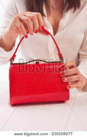 Female hands holding red handbbag. Woman with beautiful manicure holding bright lacquered clutch on white wooden table.