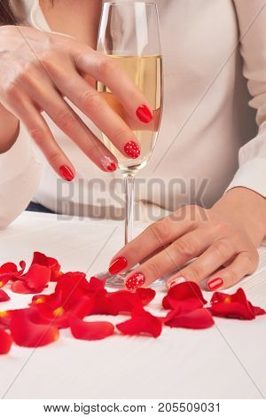 Glass of wine in female hands. Woman with beautiful manicure holding glass of wine surrounded by rose petals. Well-groomed woman hands with wine glass.