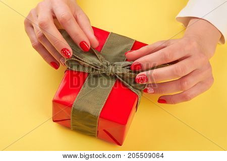 Female hands unpacking red gift box. Young woman manicured hands opening red box with present isolated on yellow background. Holiday present concept.
