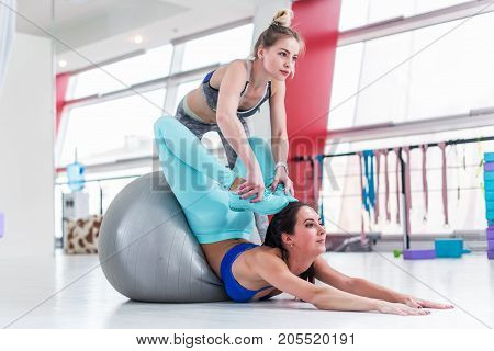 Flexible woman doing advanced stretching exercise. Pretty female instructor helping slim brunette girl do yoga Locust pose with feet touching head in gym.