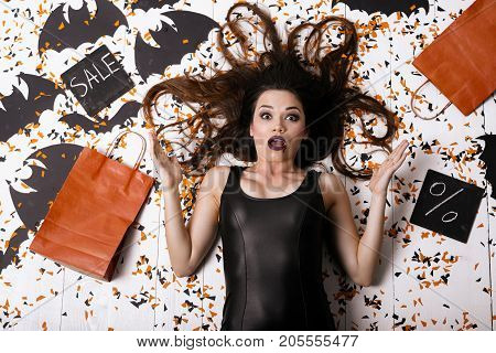 Crazy discount for Halloween. A sexy model in a black tight dress lies on the floor surrounded by confetti and paper bats. It depicts shock