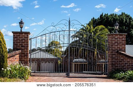 Metal driveway security entrance gates set in brick fence with residential garden in background against blue sky