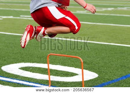A high school football player is jumping over orange mini hurdles at practice on a green turf field.