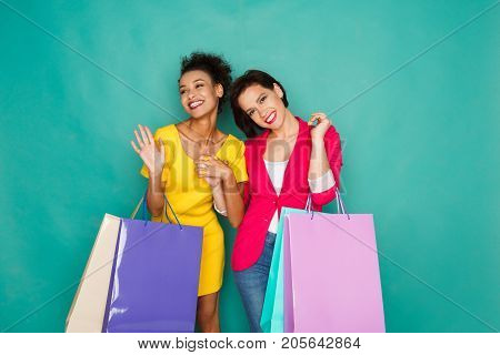Happy multiethnic girlfriends with shopping bags. Two excited shopaholics at turquoise sudio background with copy space
