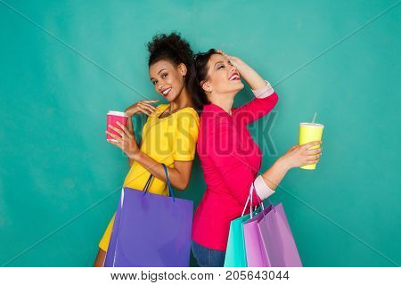 Happy multiethnic girlfriends with shopping bags and take away drinks. Two excited shopaholics at turquoise sudio background with copy space