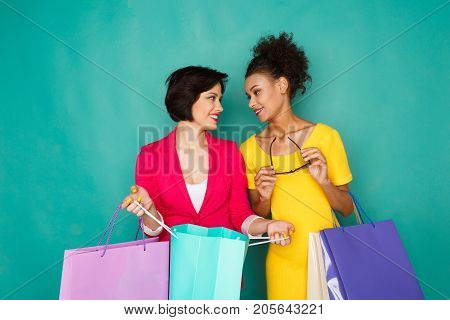 Happy multiethnic girlfriends with shopping bags. Two excited shopaholics boasting with purchases at turquoise sudio background with copy space