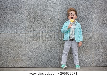 Fashion baby boy in mint jacket and sneakers stands on a gray wall background. Trendy boy with lollipop standing on the street.