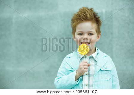 Fashion baby boy in mint jacket and sneakers stands on a gray wall background. Trendy boy with lollipop standing on the street.