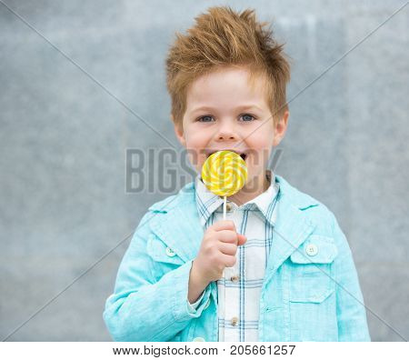 Fashion baby boy in mint jacket and sneakers stands on a gray wall background. Trendy boy with lollipop standing on the street.