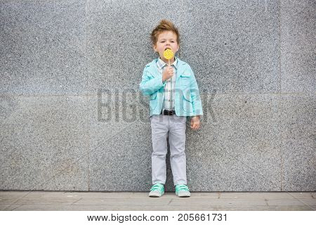 Fashion baby boy in mint jacket and sneakers stands on a gray wall background. Trendy boy with lollipop standing on the street.