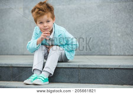 Fashion baby boy in mint jacket and sneakers posing on a gray wall background. Trendy boy standing on the street.