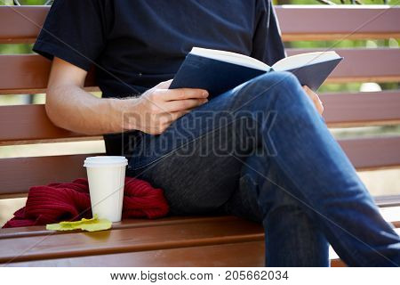 Sitting man reading a book and drinking warm tea. Cup of coffee on the bench. Close-up.
