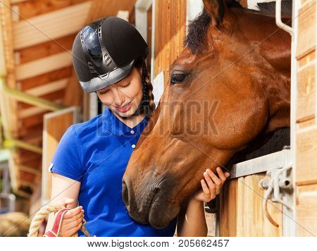 Close-up portrait of beautiful young woman in jockey skull standing by stable with her bay horse