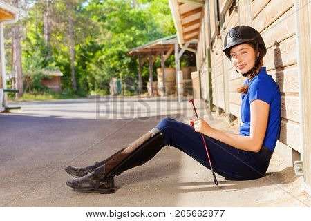 Portrait of beautiful young woman wearing jockey outfit, sitting on the floor outside a box stall and holding whip