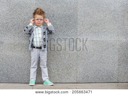 Fashion baby boy in gray jacket and mint sneakers stands on a gray wall background. Trendy boy with glasses standing on the street.
