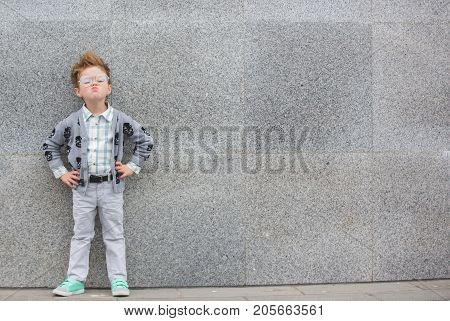 Fashion baby boy in gray jacket and mint sneakers stands on a gray wall background. Trendy boy with glasses standing on the street.