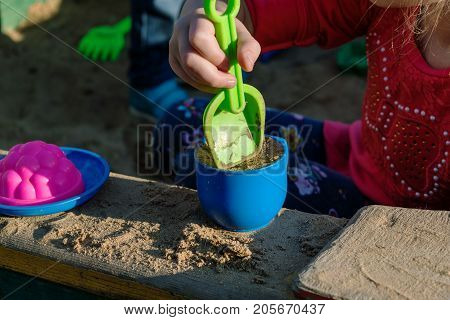 Little girl playing in a sandbox with a plastic shovel and a mug. In a mug filled with the sand lie next to a plastic plate and the mold. Summer day sun shining.