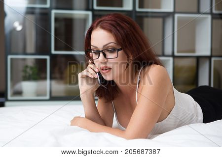 Portrait of serious sexy young Caucasian businesswoman wearing glasses lying on bed and talking on mobile phone
