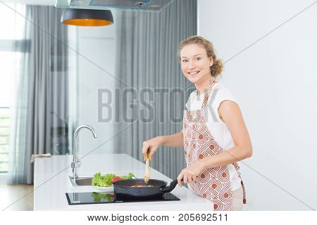Happy attractive housewife in apron preparing food at kitchen and looking at camera. Cheerful young woman stirring food in cooking pan on hotplate. Enjoying cooking concept