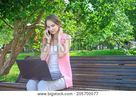 Woman sitting on bench in park talking on cell phone and using laptop