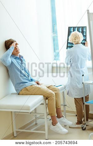 Young man sitting on a hospital daybed leaning on wall and touching his forehead with doctor examining his x-ray photograph