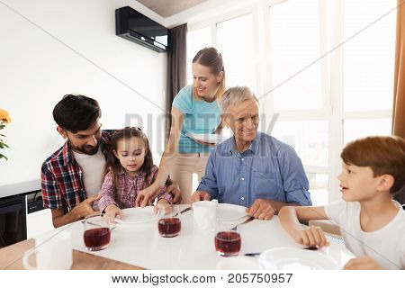 Family prepare for dinner. A man, an old man and two children are already sitting, a woman in a blue T-shirt puts the plates in front of them. Before them are already glasses of juice