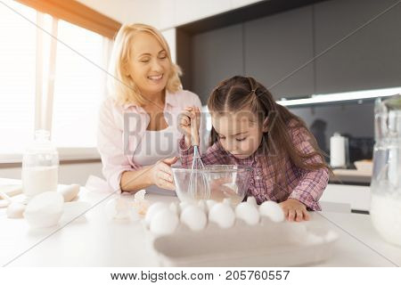 A little girl bent over a glass bowl in which she whipped eggs with a whisk. Her grandmother is standing beside her and helping her