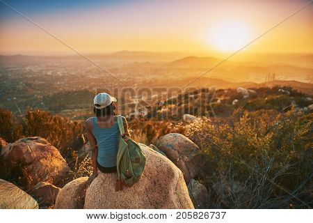 Rear view of woman hiker sitting on rock on top of hill while looking at sunset over San Diego California
